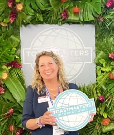 District 53 Program Quality Director Bettyann Peck, DTM, in front of the Toastmasters logo and holding a portable Toastmasters logo, at the 2023 International Convention